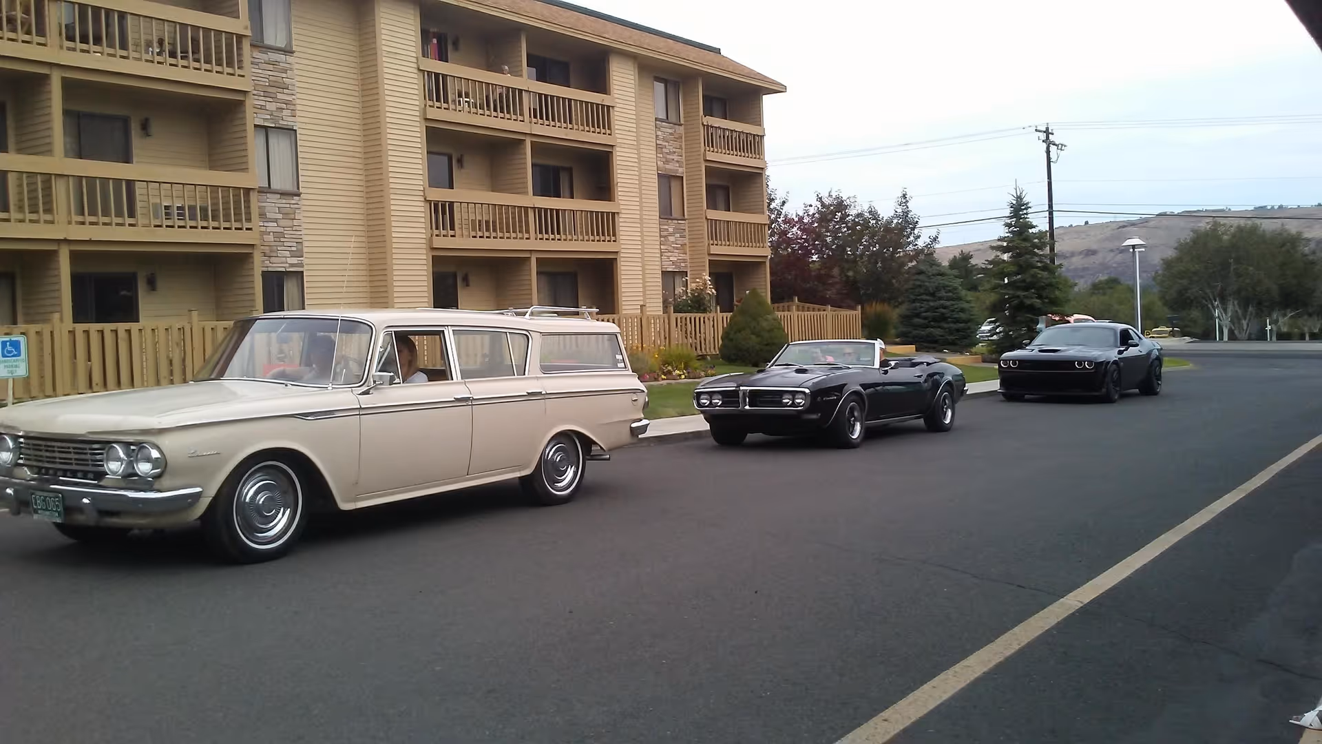 Three cars parked in a row on a paved road in front of a multi-story residential building with balconies and stone accents. The first car is a vintage beige station wagon, the second is a black convertible, and the third is a modern black sports car. There are trees and a hill in the background.