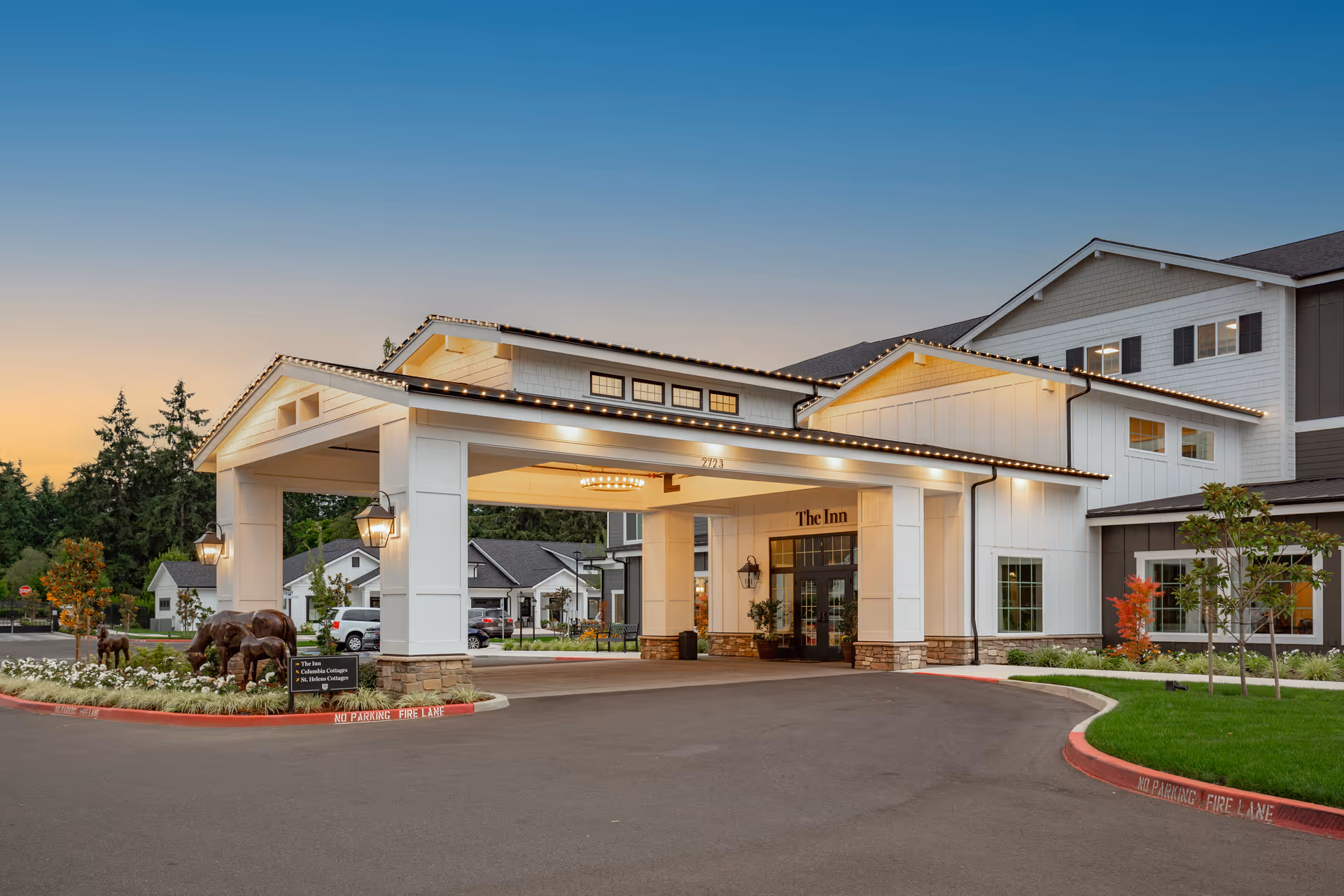 Exterior view of The Inn at University Village Assisted Living Community at dusk, showing the main entrance with a covered driveway, well-lit building facade, landscaped greenery, and a sculpture of grazing horses near the entrance.
