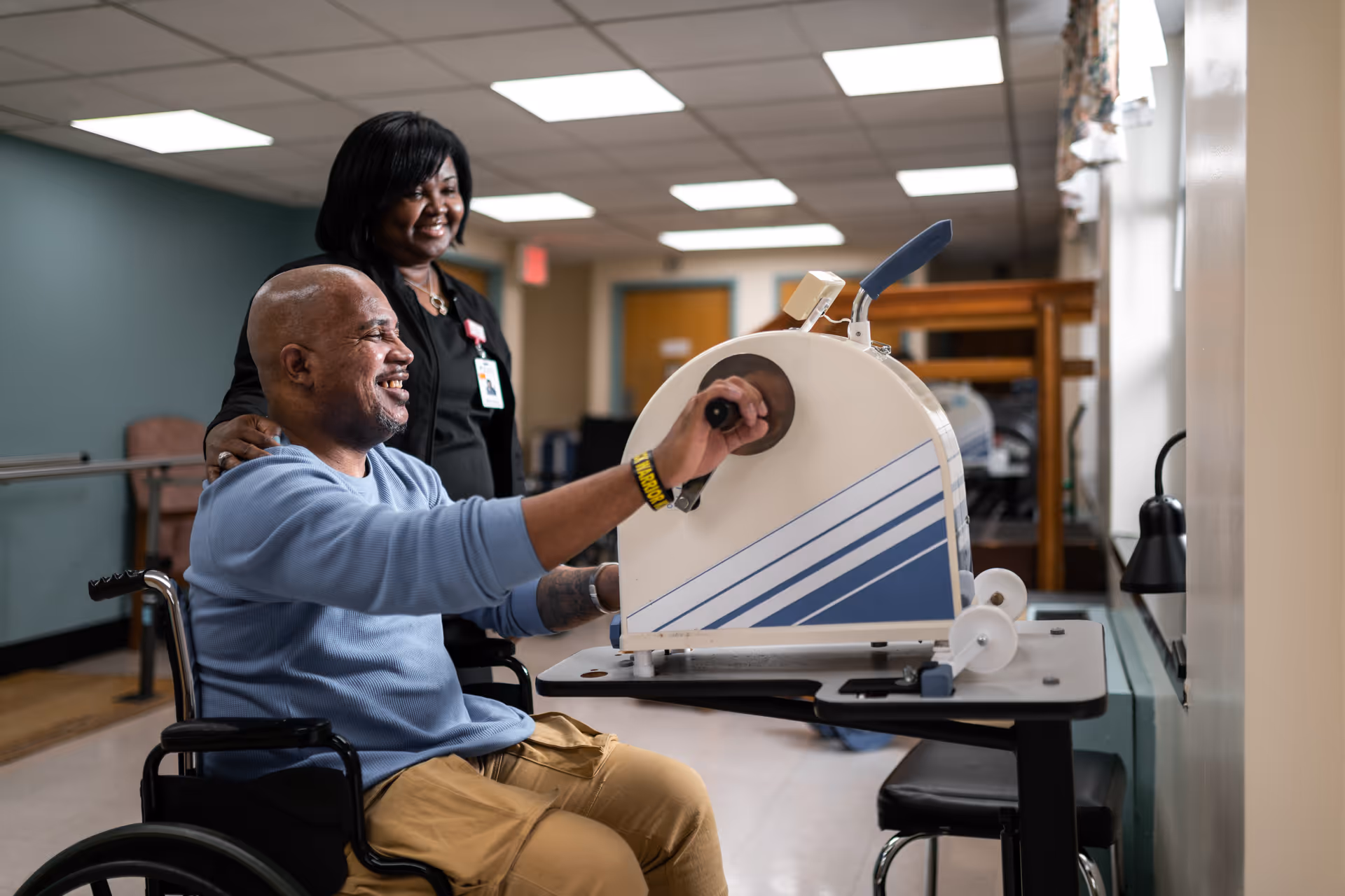 A man in a wheelchair is using a hand exercise machine in a rehabilitation room while a female staff member stands behind him, smiling and offering support.