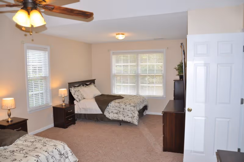 A bright bedroom with two beds featuring floral patterned bedding and dark wooden headboards. There are two nightstands with lamps, a ceiling fan with lights, a large window with white blinds, and a dresser with a plant on top. The walls are painted beige and the carpet is light brown.