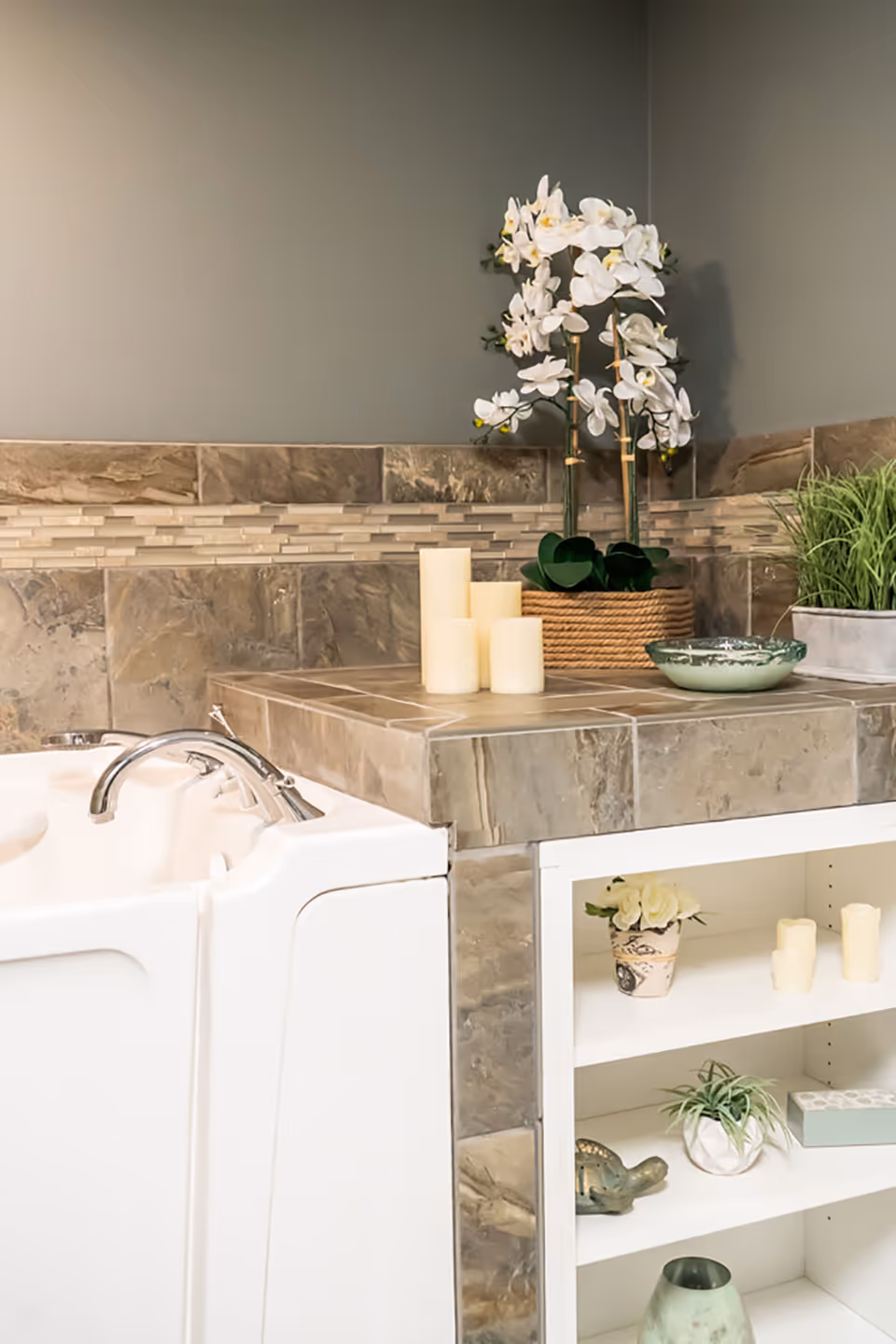 A bathroom corner featuring a white walk-in bathtub with chrome faucet, surrounded by beige and brown tiled walls and countertop. On the countertop, there are decorative items including white candles, a potted white orchid, and a green plant. Below the countertop is a white open shelf with additional decorative items such as small plants, candles, and a ceramic turtle.