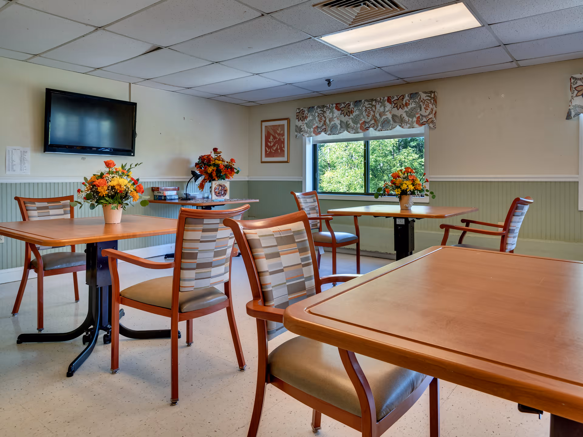 A bright dining room with several wooden tables and chairs arranged neatly. Each table has a floral centerpiece with colorful flowers. A large window with floral valances lets in natural light and offers a view of green trees outside. A flat-screen TV is mounted on the wall, and there is a framed picture hanging next to the window.