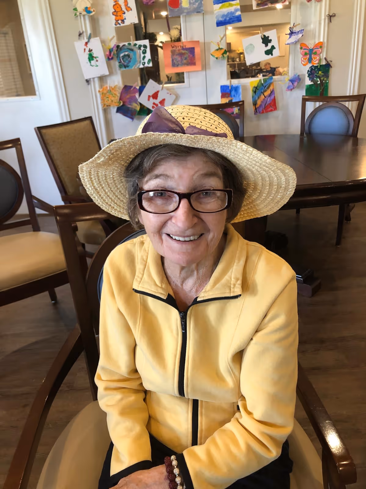 An elderly woman wearing glasses, a yellow jacket, and a straw hat with a purple ribbon is sitting on a chair in a room with wooden floors and several chairs and tables. Behind her, colorful artwork is displayed on a glass partition.