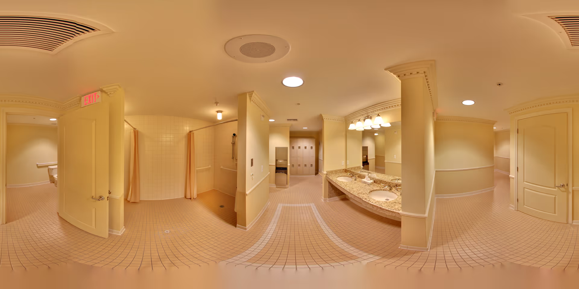 A spacious, well-lit bathroom area with beige tiled floors and walls painted in a soft yellow tone. The room features two sinks with a large mirror above them, multiple shower stalls with curtains, and several doors leading to other areas. The ceiling has recessed lighting and ventilation. There is an exit sign above one door.