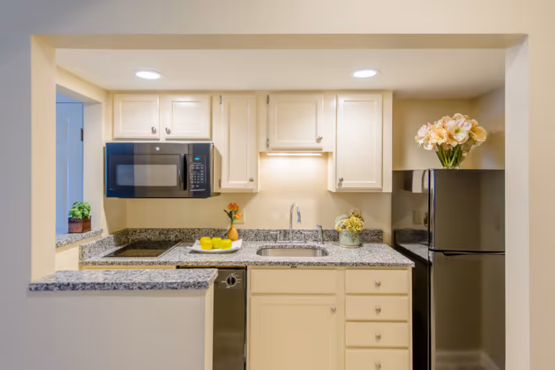 Small modern kitchen with white cabinets, granite counters, stainless appliances, and flowers on the counter.