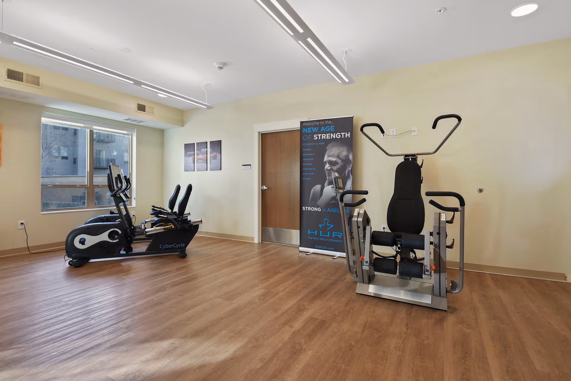 A fitness room with exercise equipment including a CyberCycle recumbent bike and a strength training machine. The room has wood flooring, a window letting in natural light, and a poster on the wall promoting strength training for seniors.