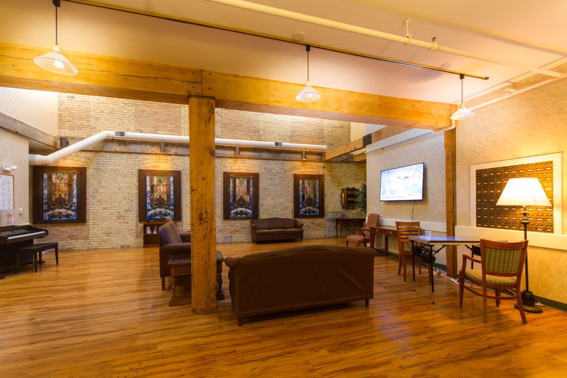 A spacious living room area with wooden floors and exposed brick walls. The room features several brown leather sofas, a piano with a bench, stained glass windows, a flat-screen TV mounted on the wall, a table with chairs, and a floor lamp providing warm lighting. Wooden beams and industrial-style pipes are visible on the ceiling.