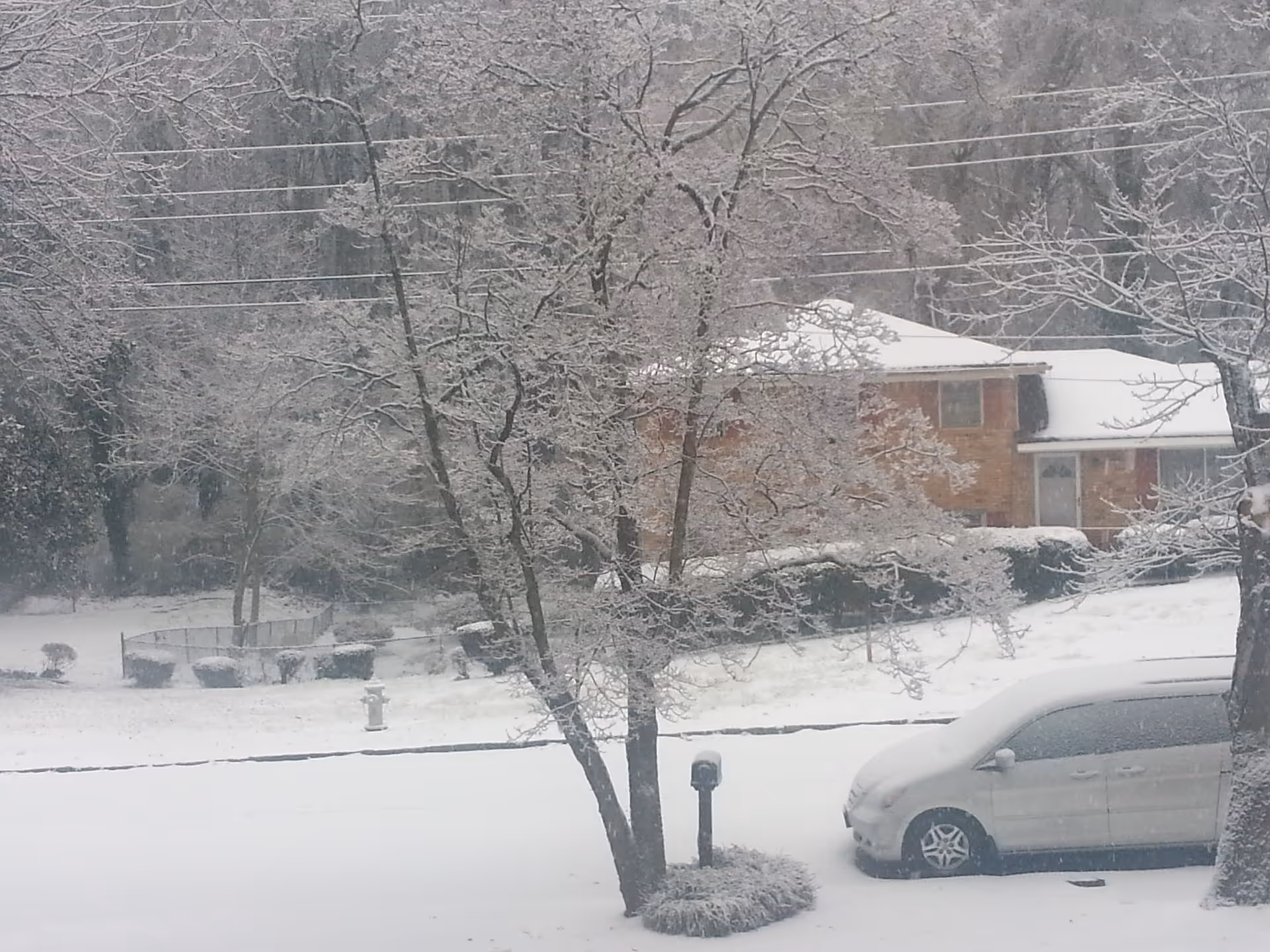 Snow-covered suburban street with a house, bare trees, and a parked minivan.