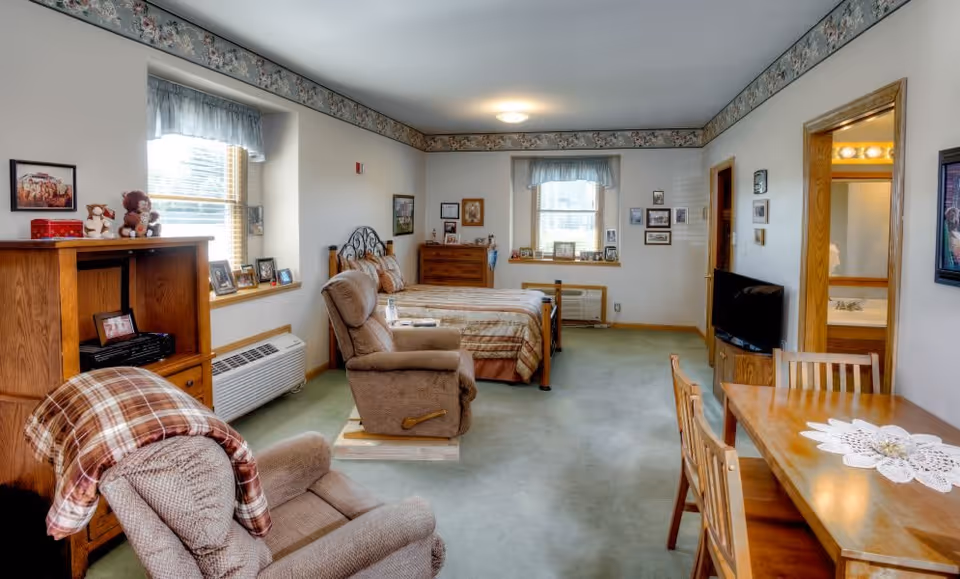A cozy senior living room and bedroom area with two recliner chairs, a wooden entertainment center, a bed with a metal headboard, a wooden dresser, a small dining table with chairs, and a television. The room has light green carpet, white walls with floral border wallpaper near the ceiling, and two windows with light blue valances. A bathroom is visible through an open door.