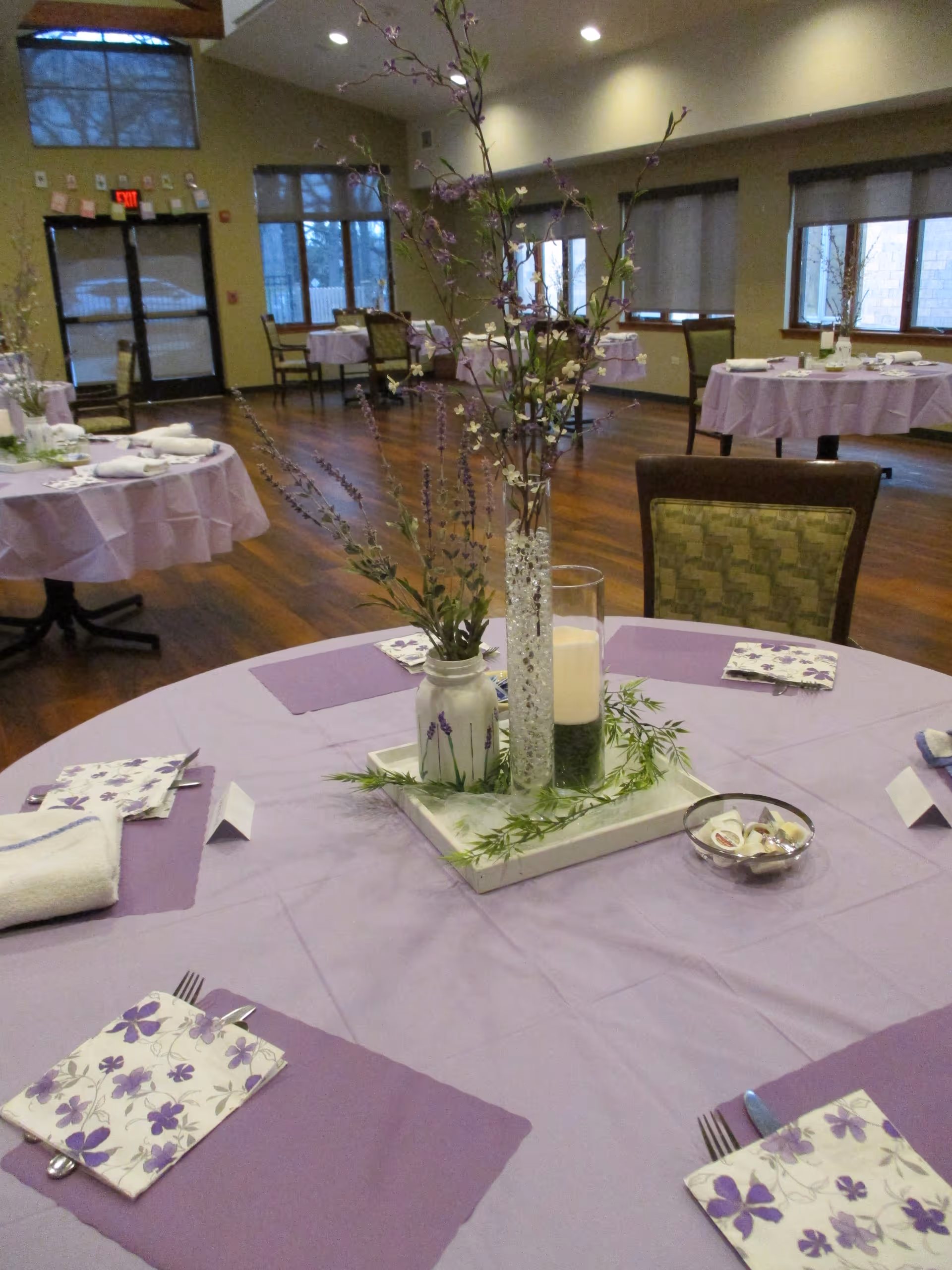 A dining room with round tables covered in light purple tablecloths and matching purple placemats. Each table is set with floral napkins, silverware, and a centerpiece featuring a tall vase with branches and flowers, a candle, and greenery. The room has wooden floors, large windows with blinds, and a glass door in the background.