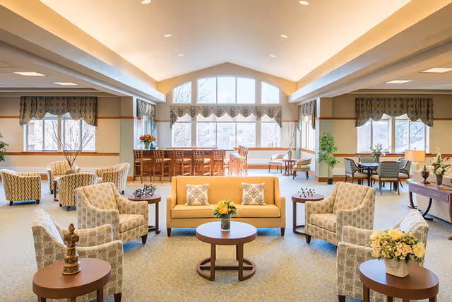 A spacious and well-lit living room area in a retirement community with multiple seating arrangements including a yellow sofa with patterned pillows, several armchairs, round wooden tables with decorative items and flowers, large windows with valances allowing natural light, and additional seating and tables in the background.