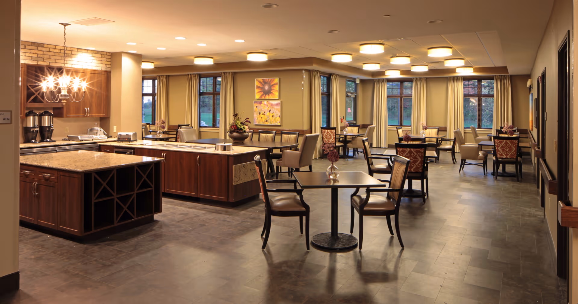 A spacious, well-lit dining room with multiple tables and chairs and a central serving island/kitchen area.