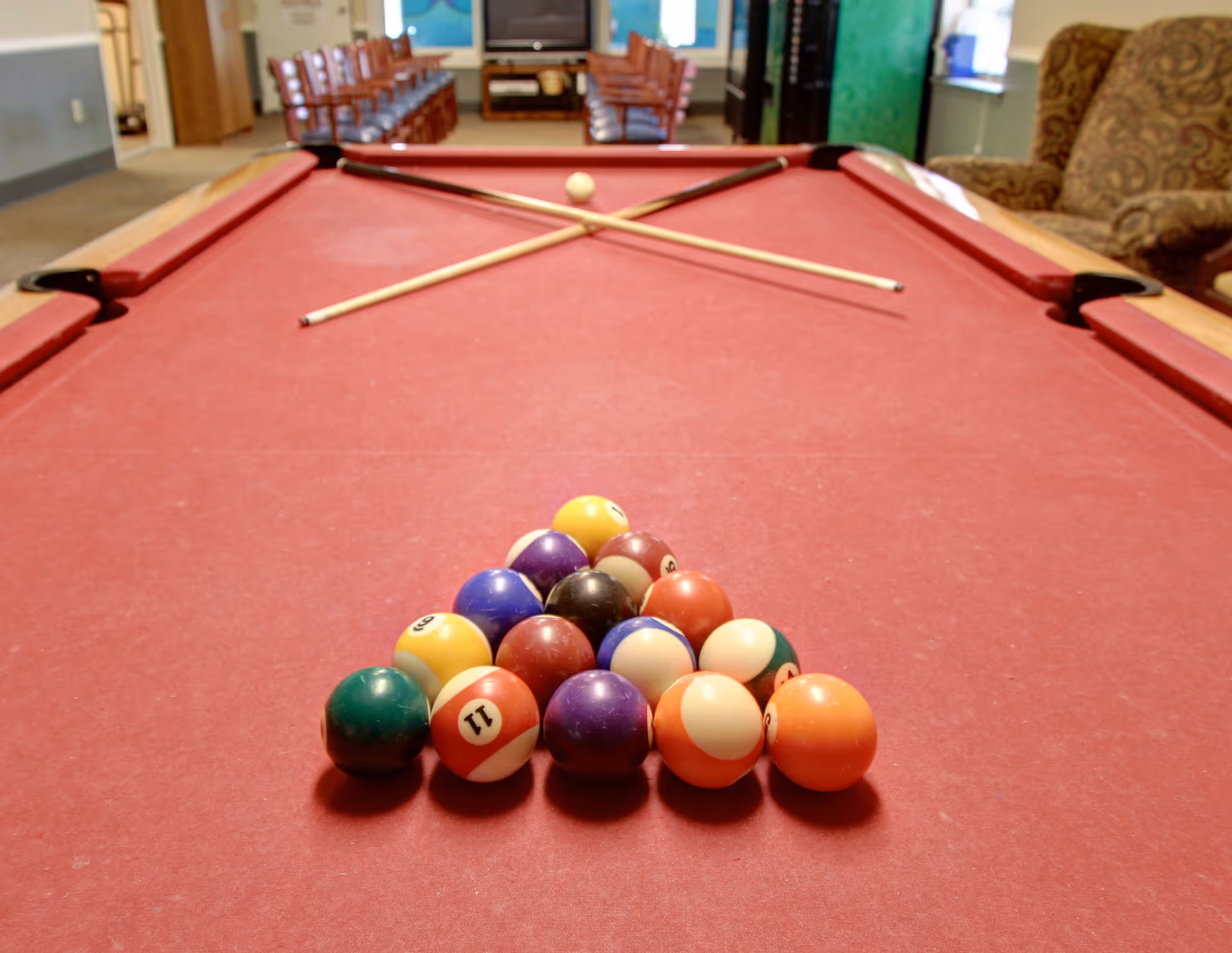 A red pool table with a full rack of billiard balls arranged in a triangle and two crossed pool cues resting on the table. In the background, there are chairs lined up against the wall and a large TV or screen mounted on the wall.
