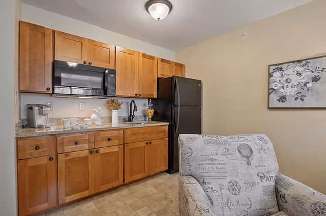 A small kitchenette with wooden cabinets, granite countertop, black microwave and refrigerator, and a patterned armchair in the foreground.