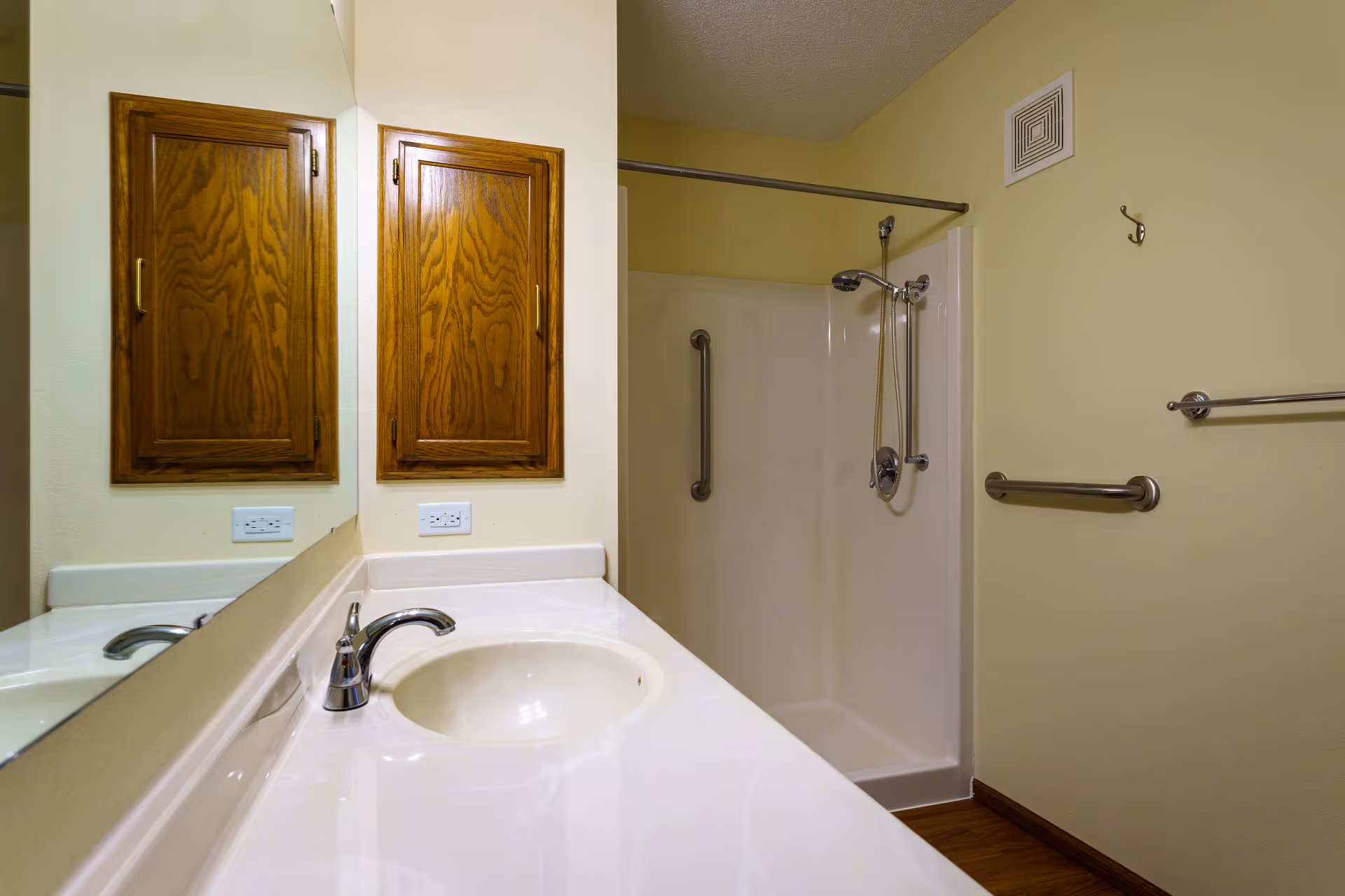 A bathroom with a white countertop and sink, a chrome faucet, a large mirror, wooden medicine cabinets, a walk-in shower with grab bars, and a showerhead with a handheld attachment. The walls are painted light yellow and there is a towel bar and a hook on the wall.