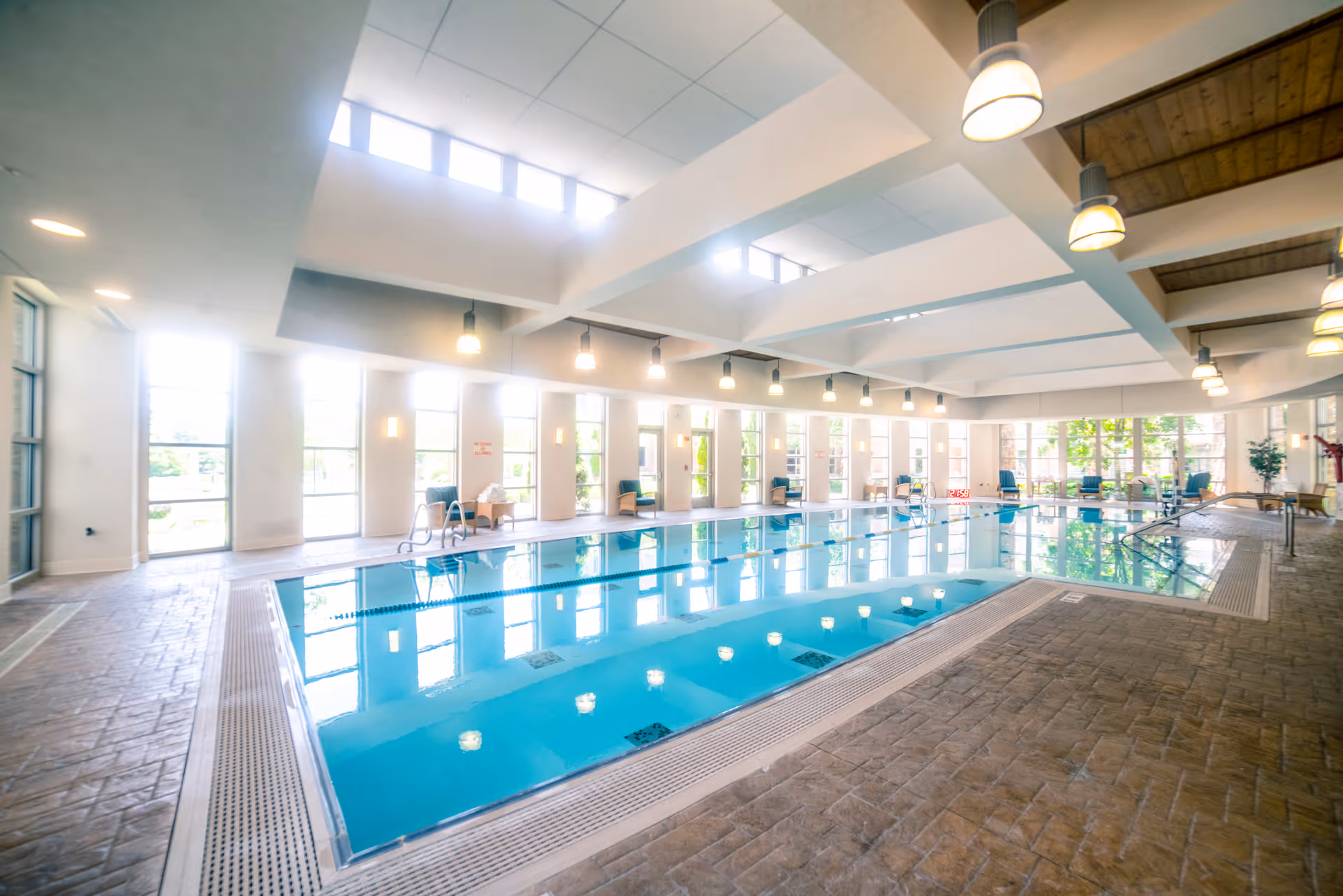 Bright indoor swimming pool area with large windows letting in natural light, several chairs along the walls, and ceiling lights illuminating the space.