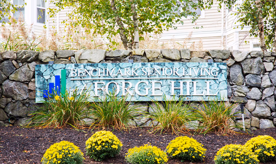 Entrance sign reading 'Benchmark Senior Living Forge Hill' mounted on a stone wall with landscaping and yellow flowers in front.