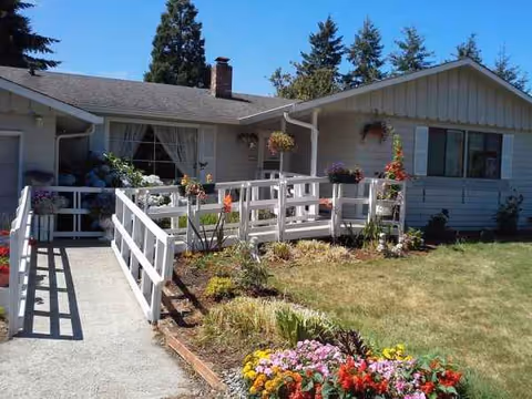 Single-story care home exterior with a white ramp, garden beds, and hanging baskets.