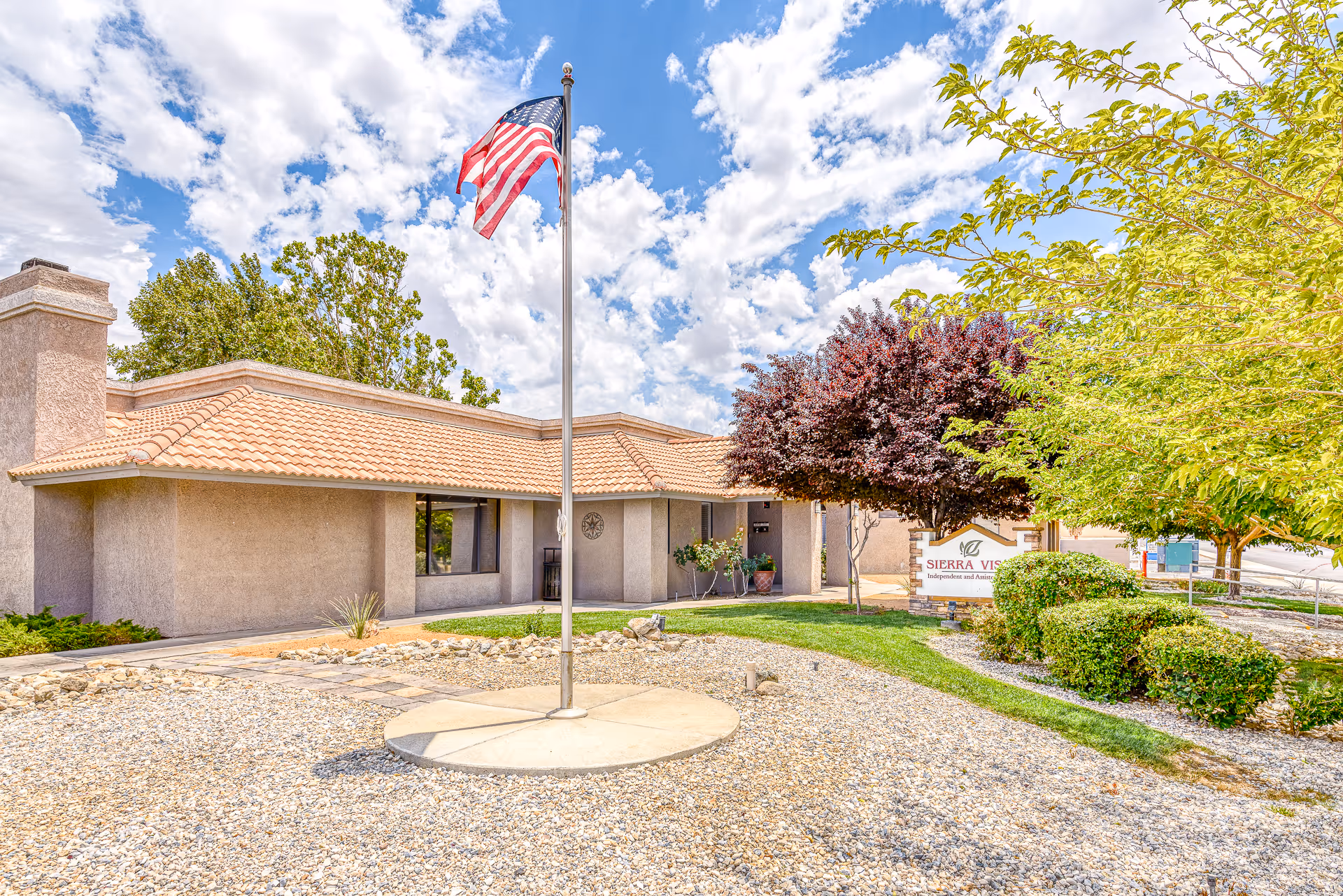 Front exterior of Sierra Vista Independent & Assisted Living showing a single-story stucco building, an American flag on a flagpole, and landscaped gravel and greenery.