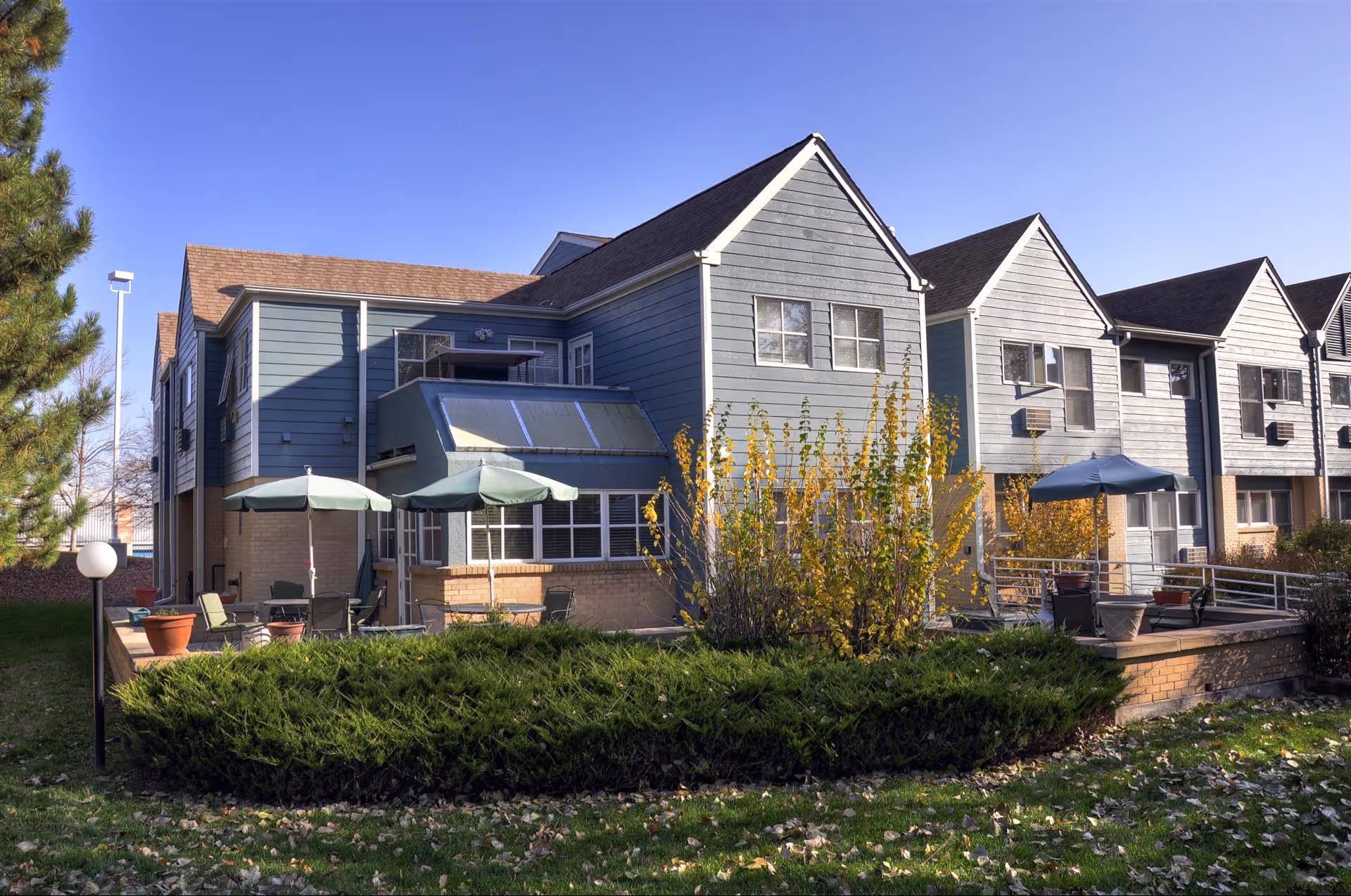 Exterior view of a two-story assisted living facility building with blue-gray siding and brown shingled roofs. The building has multiple windows and a patio area with green umbrellas, chairs, and potted plants. There are bushes and a tree in the foreground under a clear blue sky.
