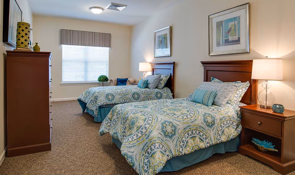 Well-lit bedroom with two twin beds dressed in blue-green patterned bedding, wooden headboards and nightstands, and a window.