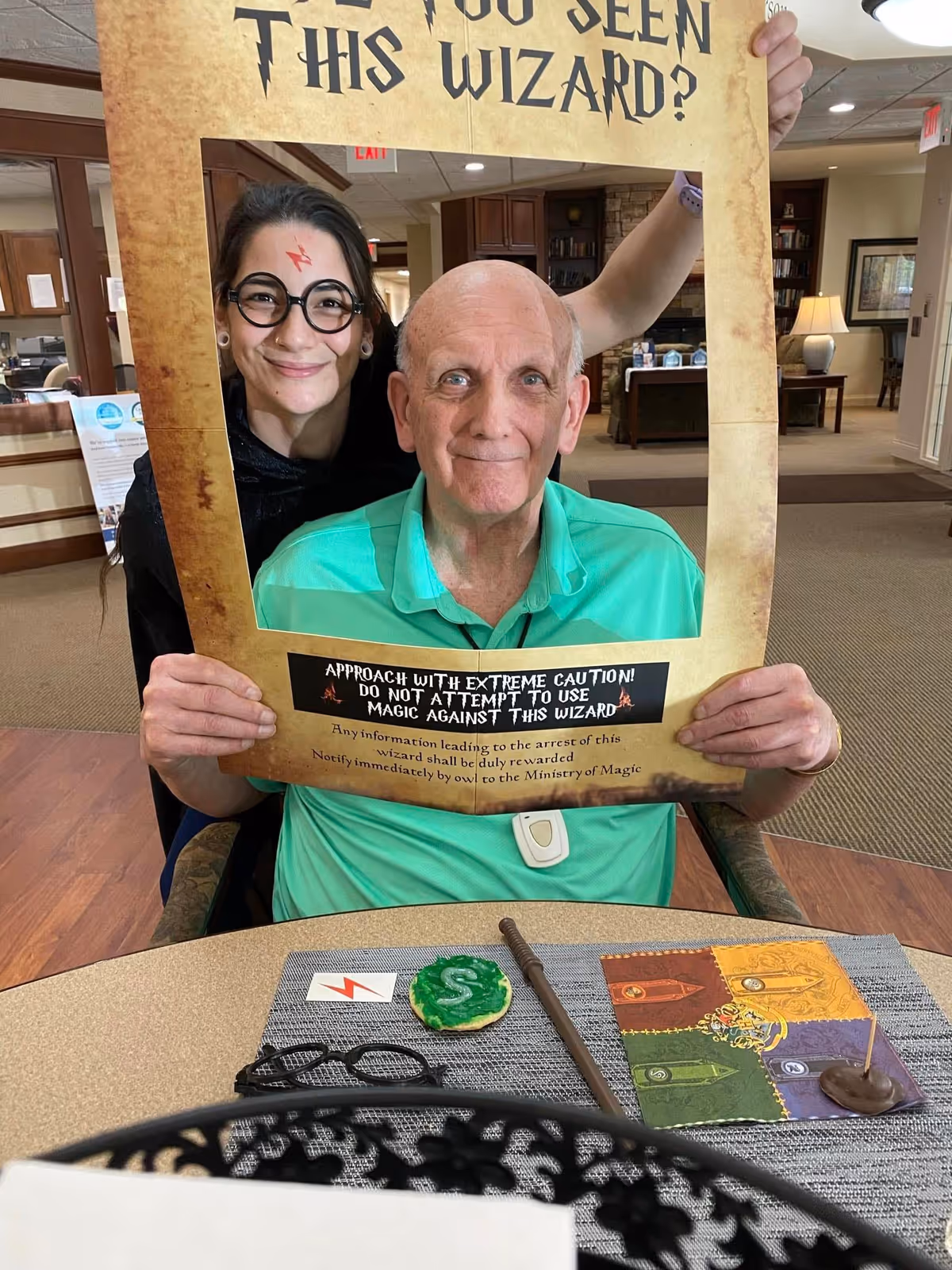 An elderly man sitting at a table holding a large cardboard frame that reads 'Have you seen this wizard?' with a warning about using magic against the wizard. A younger woman stands behind him, smiling and wearing round glasses and a lightning bolt drawn on her forehead, resembling a Harry Potter theme. On the table are themed items including a wand, glasses, a cookie decorated with a green 'S', and a colorful cloth with house crests.