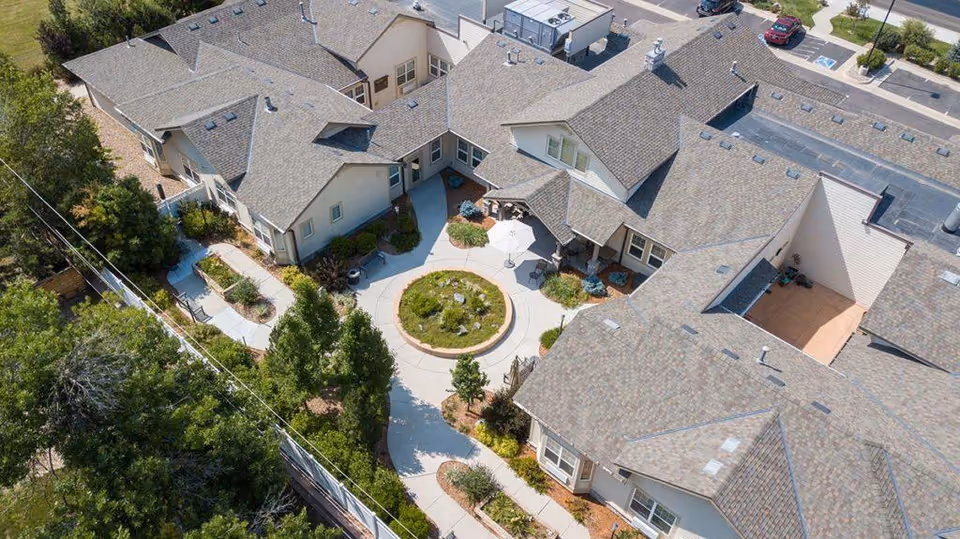 Aerial view of a senior living facility named Gardens at Columbine showing multiple connected buildings with gray shingled roofs surrounding a circular courtyard with a small garden and umbrella-covered seating area. Sidewalks and landscaped greenery are visible around the buildings.