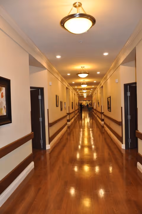 A long, well-lit hallway in a nursing and rehabilitation facility with polished wooden floors, beige walls, handrails on both sides, framed artwork on the walls, and multiple open doorways leading to rooms.