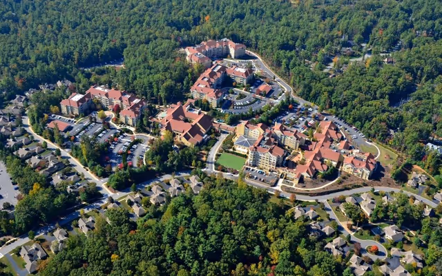 Aerial view of Deerfield Episcopal Retirement Community showing multiple large buildings with red roofs surrounded by dense green trees and smaller residential houses nearby. Parking lots with cars are visible around the buildings.