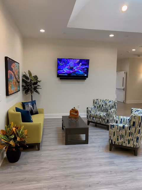 A cozy living room area with a green sofa, two patterned armchairs, a dark wooden coffee table, and a flat-screen TV mounted on a white wall displaying an underwater scene. There are two potted plants, one near the sofa and one on the floor, and a colorful painting on the wall beside the sofa. The room has light wood flooring and recessed ceiling lights.