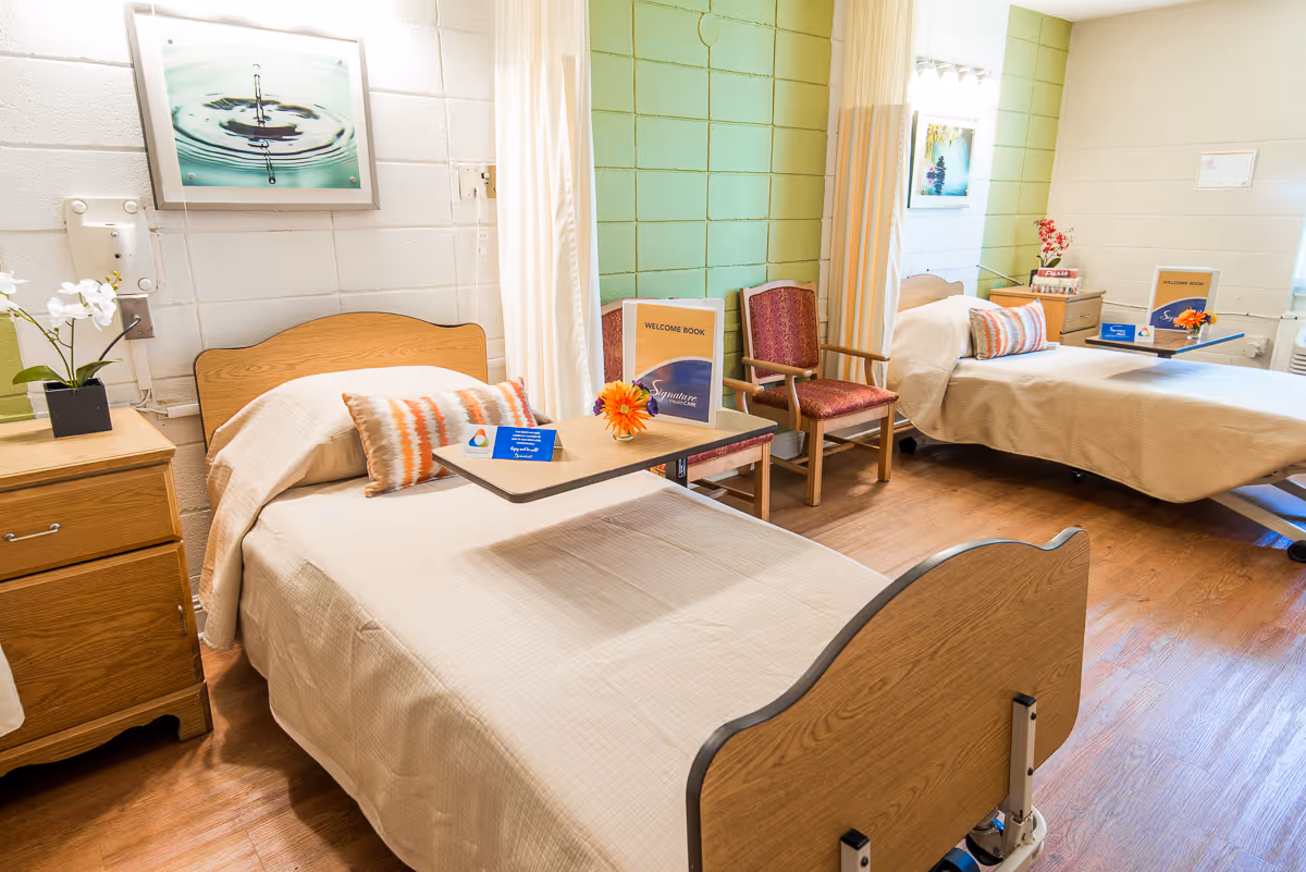 A bright and clean room with two hospital-style beds, each with a beige bedspread and a decorative pillow. Each bed has an overbed table with a welcome book and a small flower arrangement. The room has wooden flooring, light green and white painted cinder block walls, two wooden chairs with red upholstery, a wooden nightstand with a white orchid plant, and a framed picture of a water droplet on the wall.