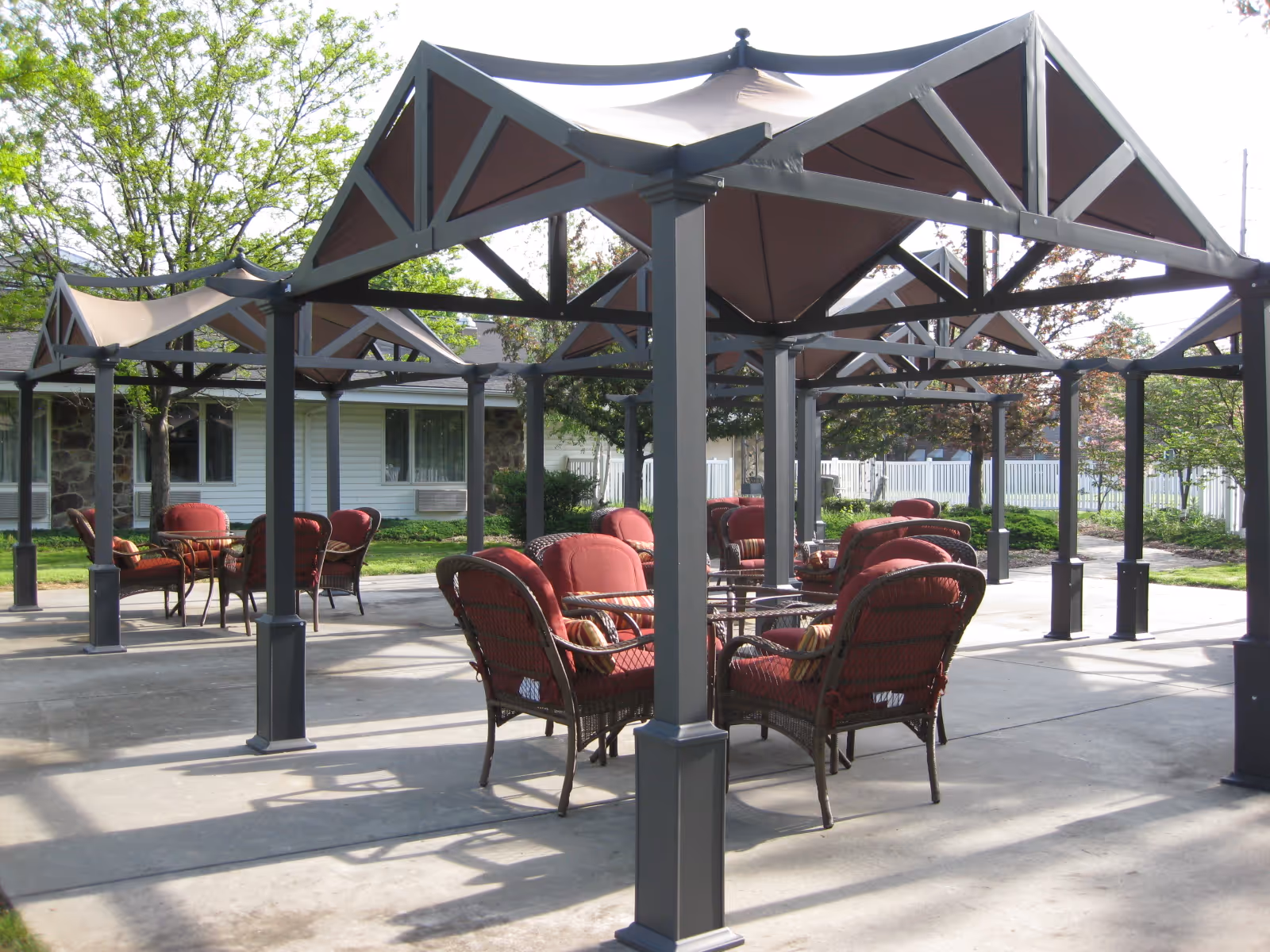 Outdoor patio area featuring multiple metal canopies with red-cushioned wicker chairs and tables beside a single-story building.