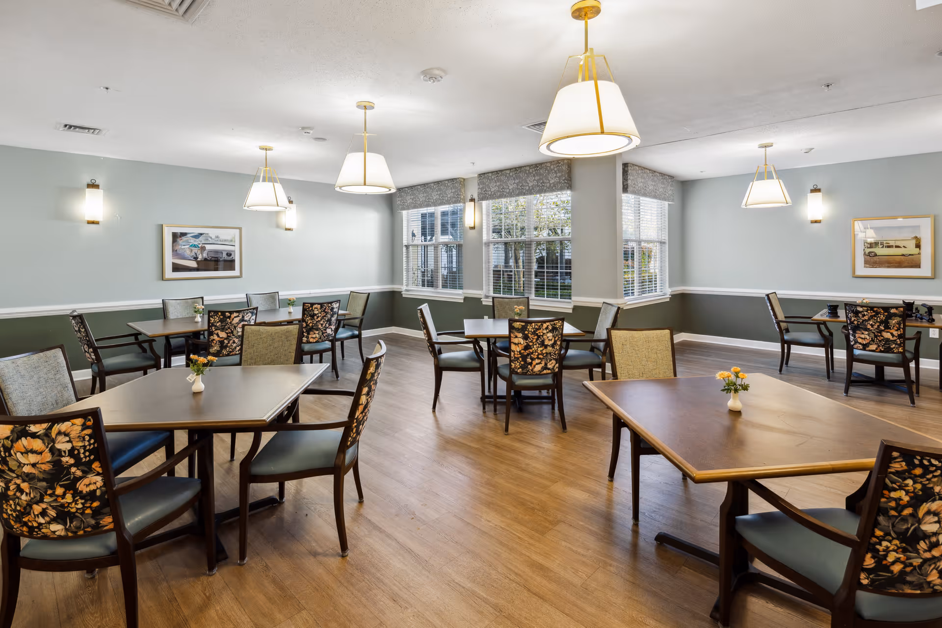 Bright communal dining room with multiple tables, floral-upholstered chairs, pendant lights, and large windows.