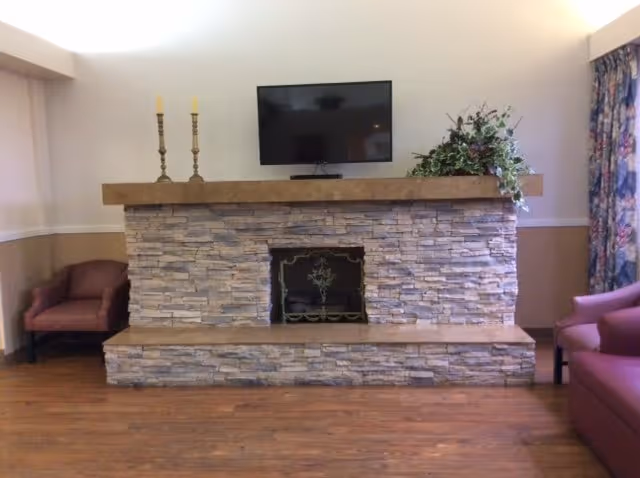 A cozy living room area featuring a stone fireplace with a wooden mantel. On the mantel, there are two tall candlesticks and a flat-screen TV mounted above. To the right of the TV, there is a green leafy plant. The room has wooden flooring and is furnished with two purple upholstered chairs, one on each side of the fireplace. A window with floral curtains is visible on the right side.