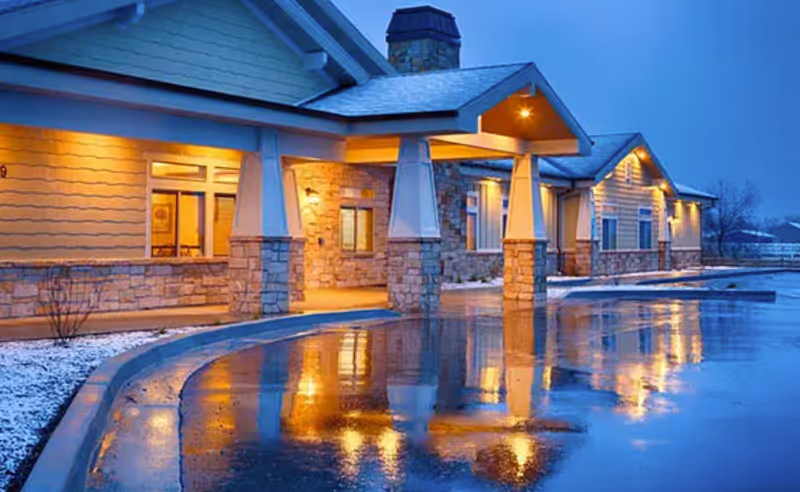Exterior view of a single-story assisted living facility building at dusk with warm lights illuminating the stone and siding facade, a covered entrance, and a wet driveway reflecting the lights.