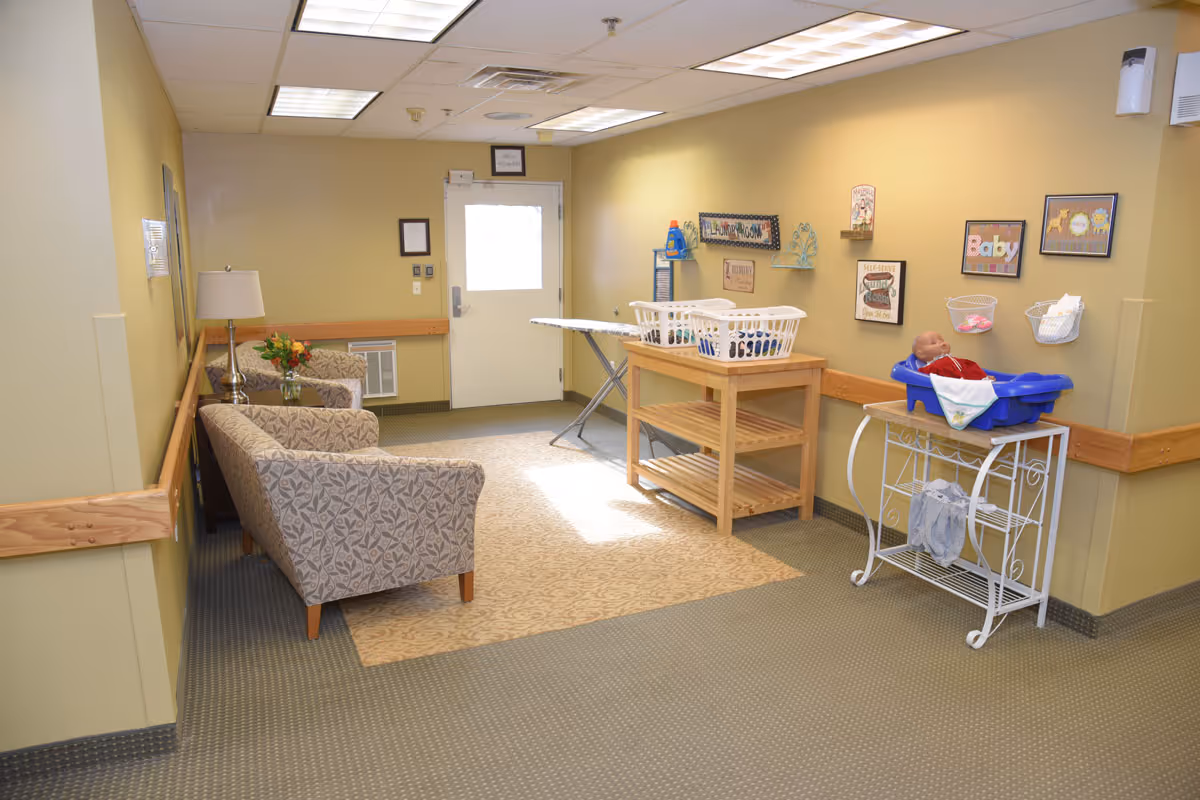 A well-lit laundry room in a senior living facility with beige walls and carpeted floor. The room features two patterned armchairs with a small table and lamp between them on the left side. On the right side, there is a wooden folding table with laundry baskets and an ironing board. A baby doll is placed in a blue baby bathtub on a white metal stand. Various decorative signs and framed pictures are hung on the wall above the laundry area.