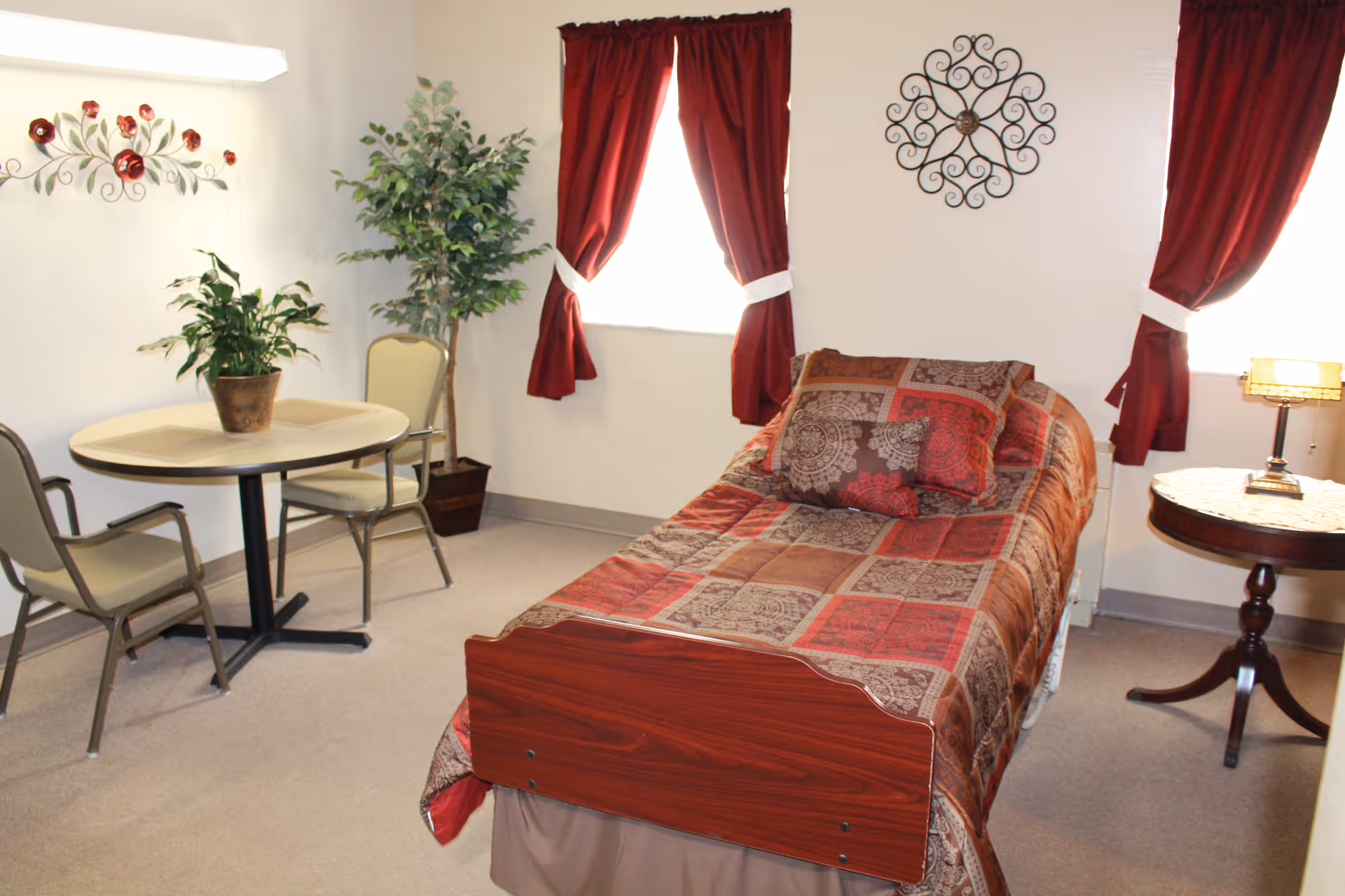 A furnished resident bedroom with a single bed, patterned bedding, red curtains, a round table with chairs, and potted plants.