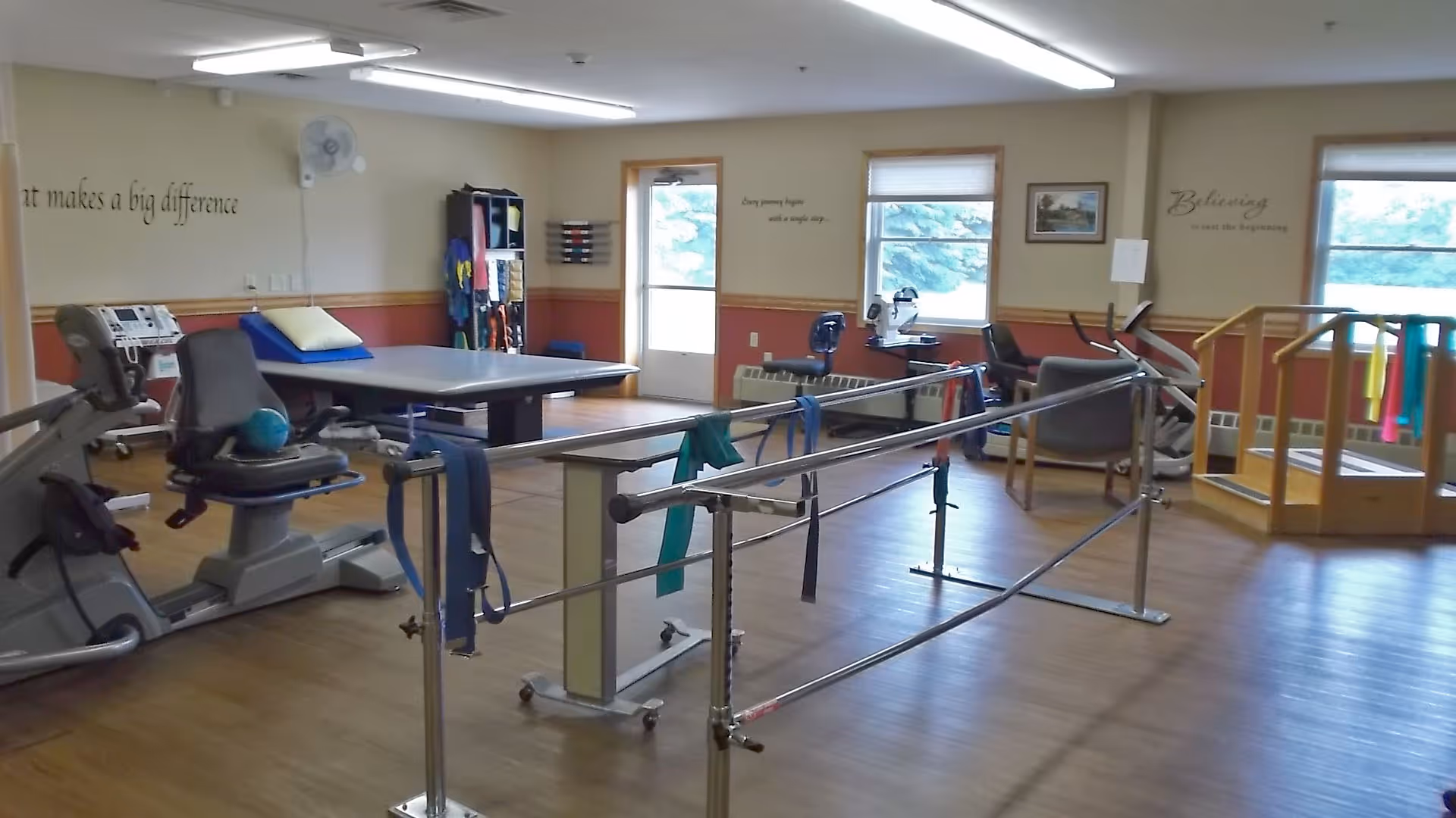 A rehabilitation or physical therapy room with exercise equipment including a recumbent bike, parallel bars with resistance bands, a padded therapy table, and stairs with handrails. The room has wooden flooring, windows letting in natural light, and motivational quotes on the walls.
