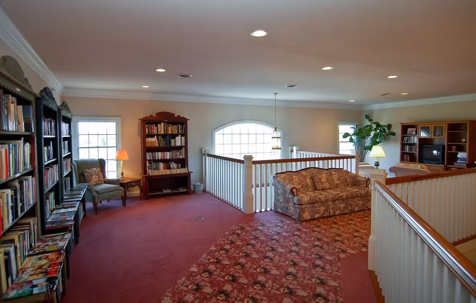 A cozy interior common area with bookshelves filled with books along the left wall, a green armchair with a side table and lamp, a floral patterned sofa, a large potted plant, and a wooden entertainment center with a TV. The room has red carpet with a floral pattern, white railings around a stairwell, and multiple windows letting in natural light.