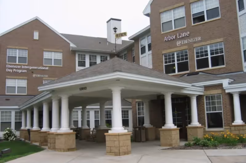 Exterior view of a senior living facility building with a covered entrance supported by white columns. The building has multiple windows and signs that read 'Ebenezer Intergenerational Day Program' and 'Arbor Lane Ebenezer'. There are some plants and flowers near the entrance.