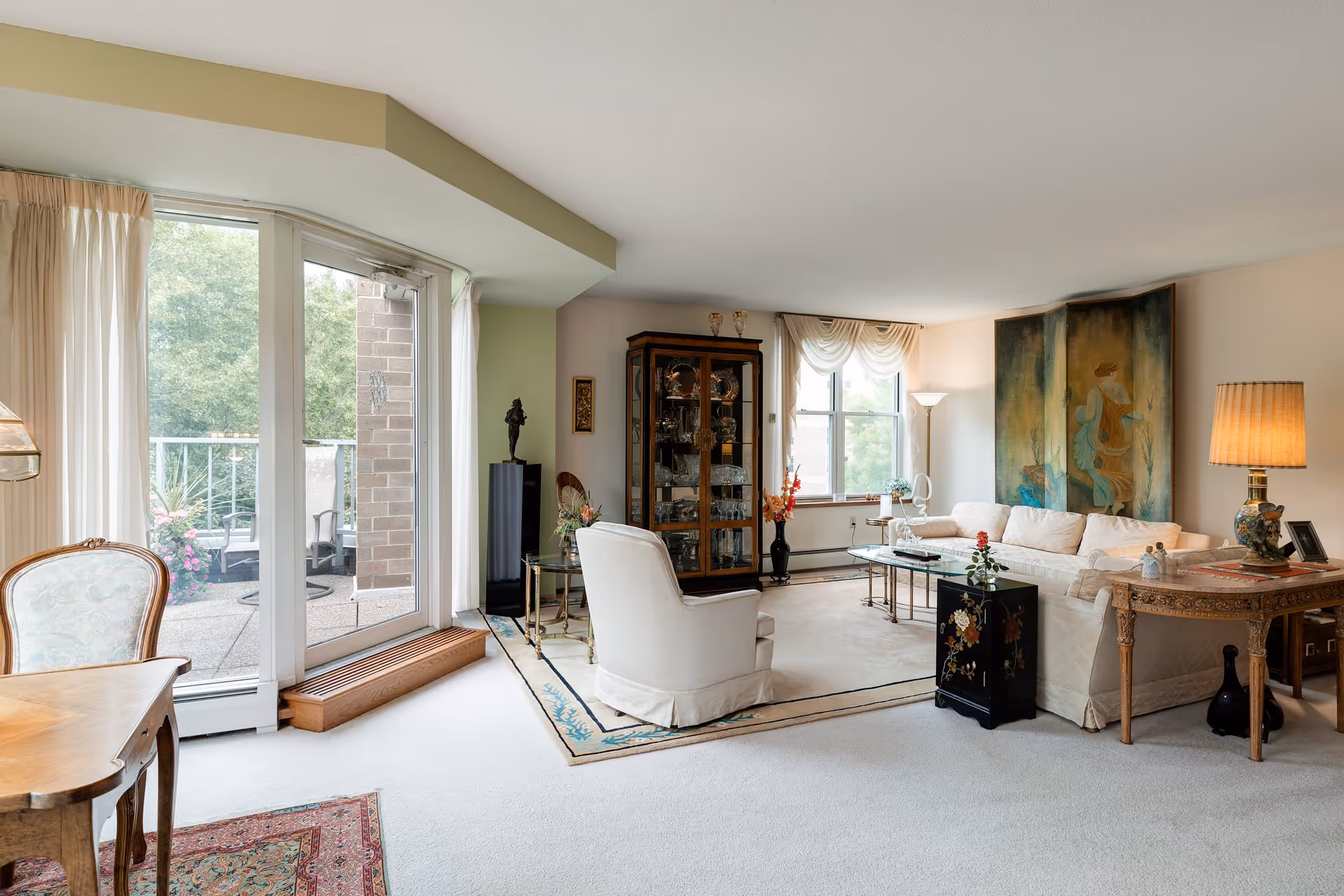 A bright and spacious living room with a large window and glass door leading to an outdoor patio. The room features a white sofa, a white armchair, a glass coffee table, a wooden side table with a lamp, a glass display cabinet, and decorative artwork on the walls. Light-colored carpet covers the floor, and sheer curtains frame the windows and door.
