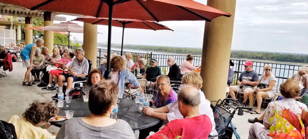 A group of elderly people sitting and socializing at round tables with umbrellas on a spacious outdoor patio overlooking a river. The patio has large columns and a railing along the edge, with greenery and trees visible in the distance.