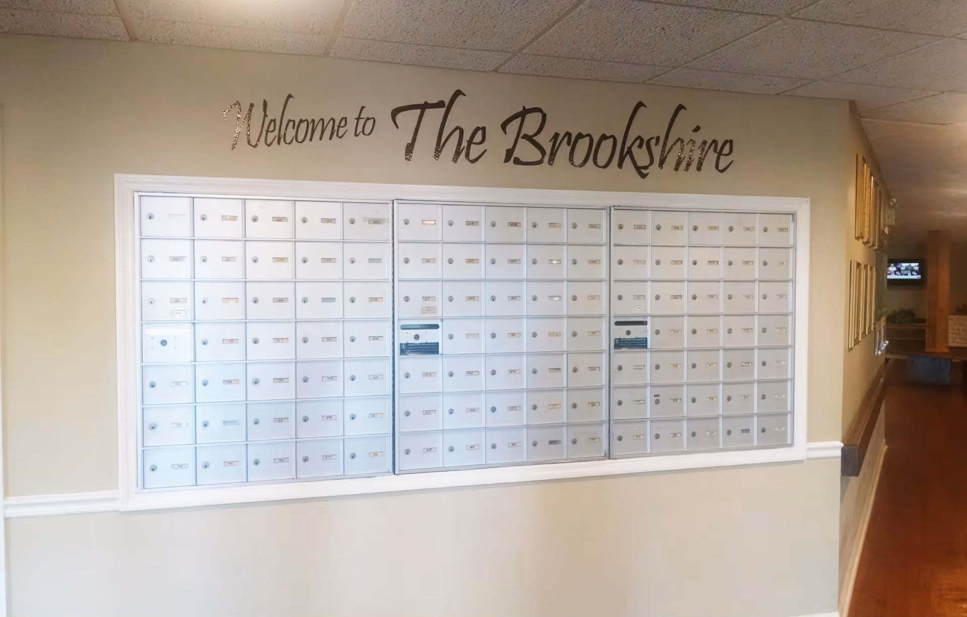 A wall with multiple white mailboxes arranged in a grid, with a sign above that reads 'Welcome to The Brookshire'. The area has beige walls, a white chair rail, and a hallway extending to the right with framed pictures on the wall and a television in the distance.