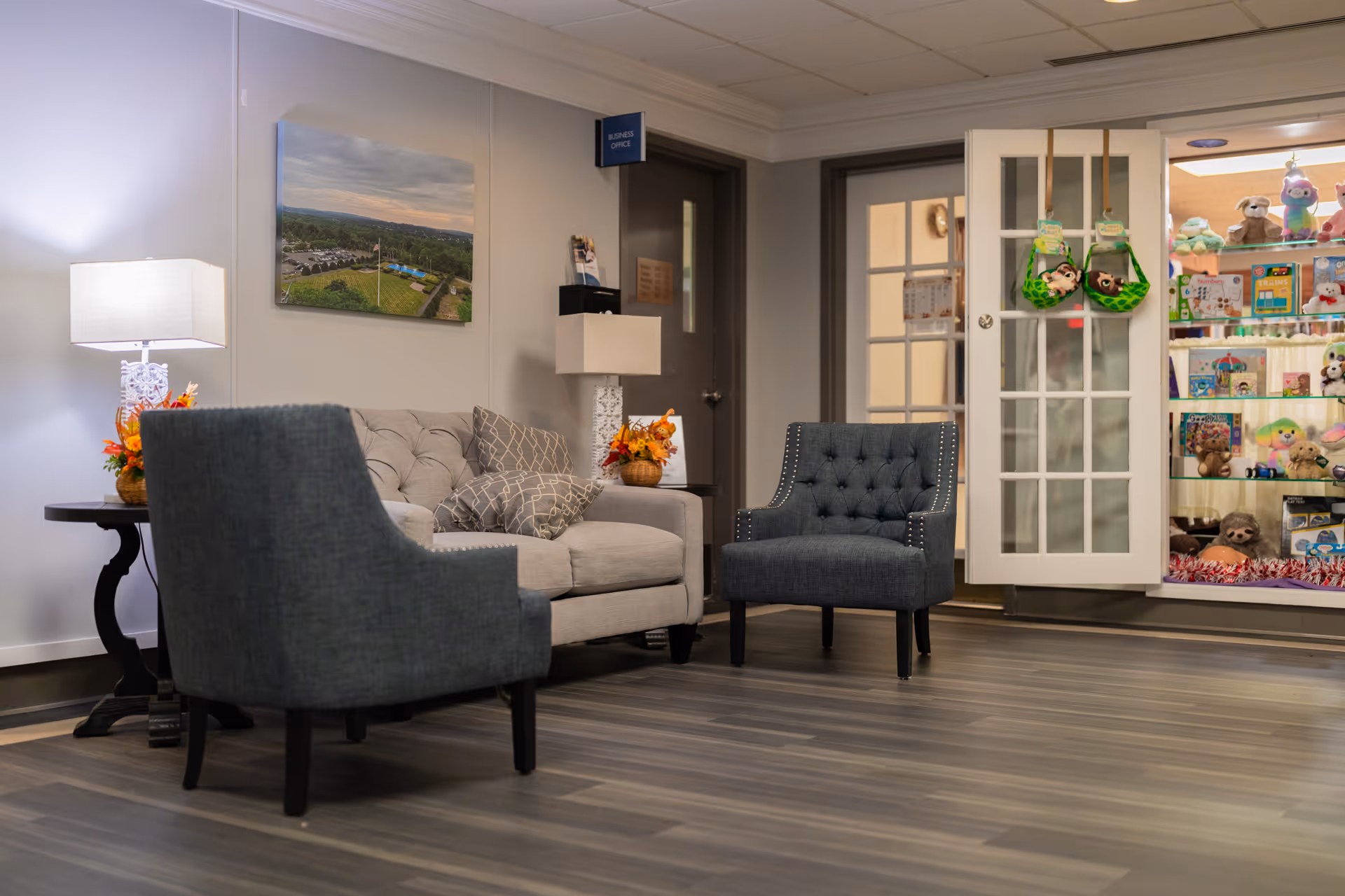 A cozy interior lounge area with a gray tufted sofa and two dark gray armchairs on a wood floor. There are two table lamps with white shades on either side of the sofa, and a framed aerial photograph on the wall. To the right, there is a glass display cabinet filled with stuffed animals and toys. A door labeled 'Business Office' is visible in the background.
