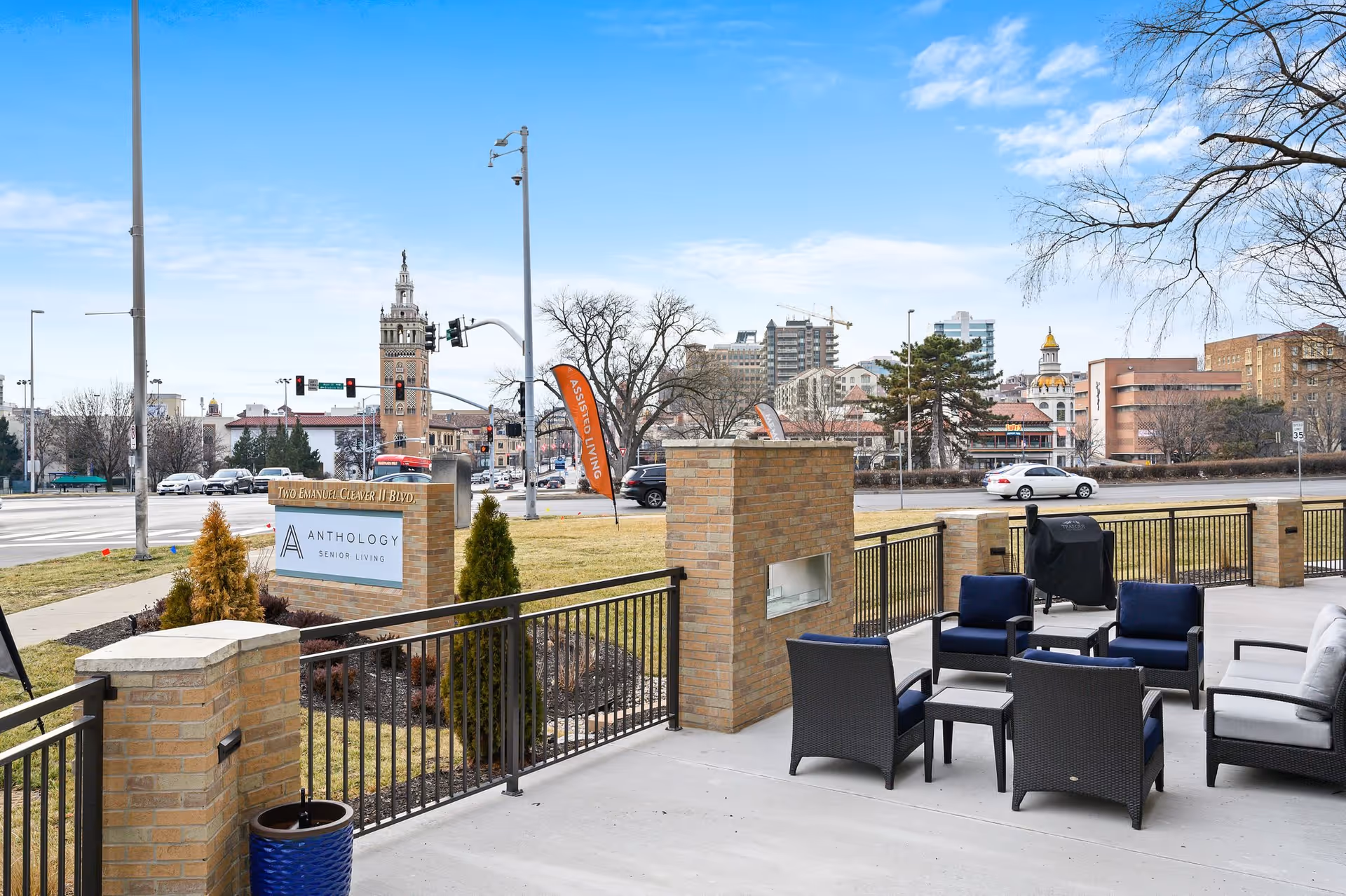Outdoor patio area with modern seating including chairs and a sofa arranged around a small table and a built-in brick fireplace. The patio is enclosed by a black metal railing and overlooks a street with buildings and trees in the background under a partly cloudy sky.