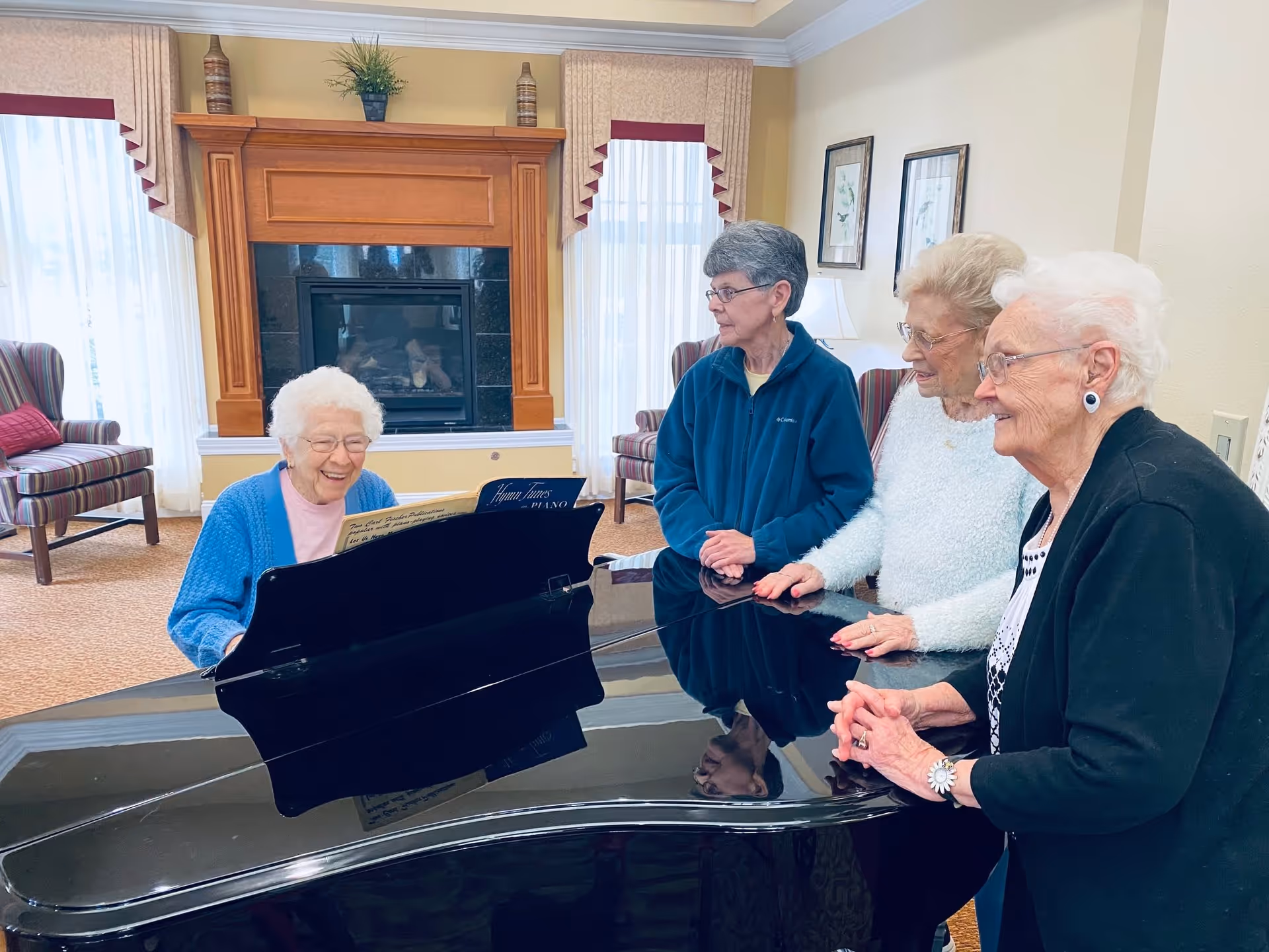 Four elderly women gathered around a black grand piano in a cozy room with a fireplace and large windows with curtains. One woman is seated playing the piano and smiling, while the other three women stand nearby watching and smiling.