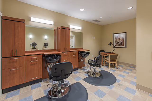 Interior view of a hair salon area in a senior living facility with two black salon chairs in front of sinks and wooden cabinets with mirrors. There is a small table with two chairs and a desk lamp in the corner, and a framed picture on the wall.