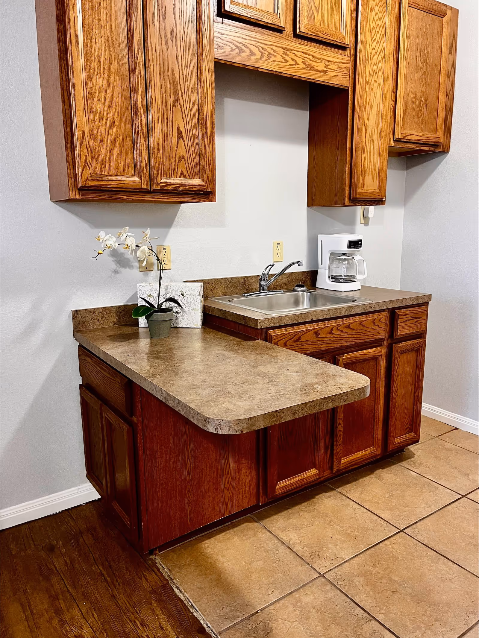 A small kitchen area with wooden cabinets, a countertop, a stainless steel sink, a white coffee maker, and a small potted orchid plant. The floor has a combination of wood and tile.