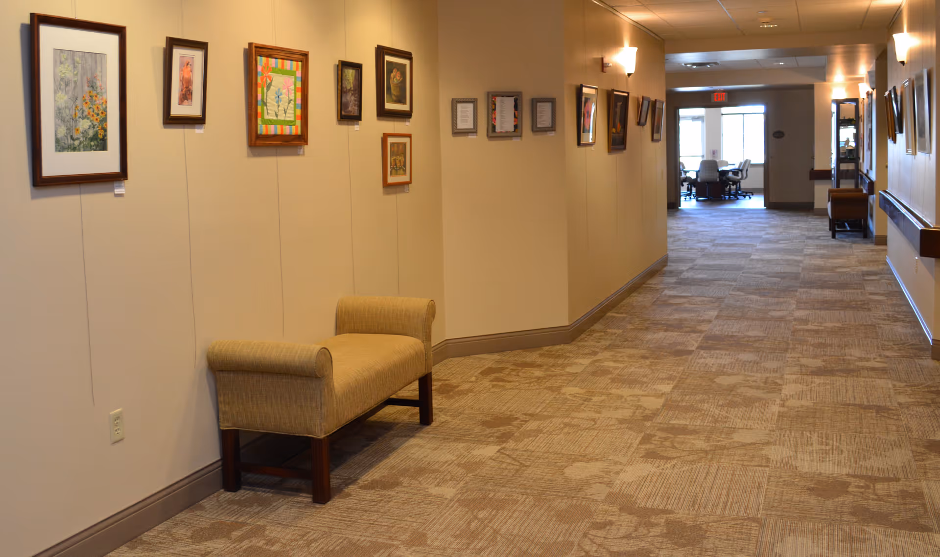 Carpeted interior hallway with framed artwork on the walls, a bench along the left wall, and a seating area visible at the far end.