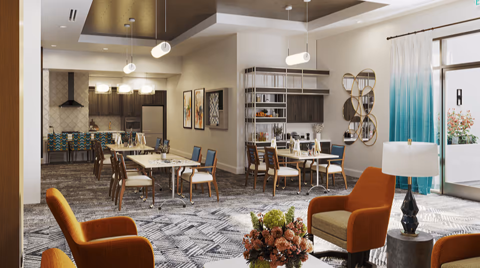 Communal dining and lounge area with dining tables, a kitchenette, and orange armchairs in a bright assisted-living interior.